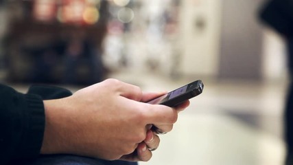 Wall Mural - An impressive closeup of young man`s hands keeping a smartphone and scrolling the information while sitting in an airy shopping mall