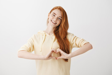 Wall Mural - Woman expresses her affection and tender feelings. Good-looking redhead woman in trendy hoodie showing heart gesture over chest, smiling broadly, confessing in love, being happy and romantic