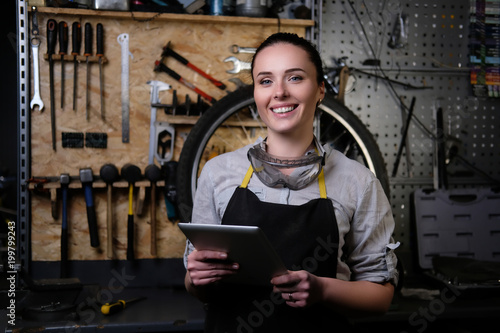 Fotografía Portrait of a beautiful brunette female wearing working clothes, apron and goggles, holds a tablet computer in a workshop