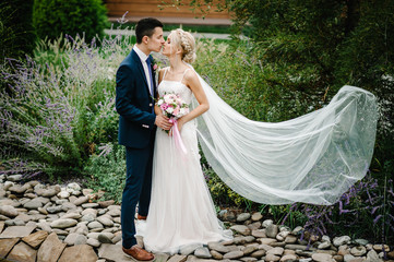 Wall Mural - Portrait of an attractive bride who embraces and kissing the groom standing and holding bouquet of pink and purple flowers and greens with ribbon. Wedding ceremony on nature.