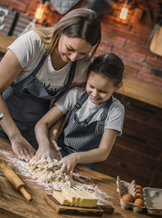 Poster - Cheerful woman and daughter in aprons preparing dough on kitchen at home together. 