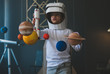 Cute little boy wearing cardboard astronaut helmet flying toy rocket through planets, cardboard spaceship rocket in the background