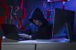 © semenyaka - A hacker in a hood sits in a dark room with a black laptop