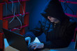 © semenyaka - A hacker in a hood sits in a dark room with a black laptop