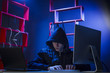 © semenyaka - A hacker in a hood sits in a dark room with a black laptop
