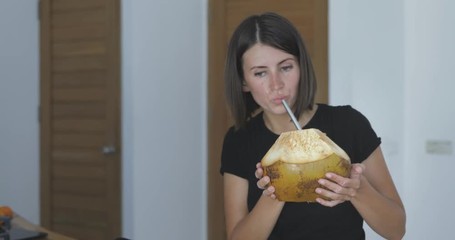 Wall Mural - Young woman drinks coconut water through reusable metal straw and eats banana. Close-up of fruits lie in string shopping bag on kitchen table . Zero waste concept. 4k video shooting by handheld gimbal