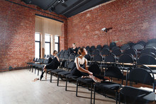 The Ballet Concept. Young Ballerina Girls Sitting On The Black Chairs In The Hall. Women At The Rehearsal In Black Bodysuits. Prepare A Theatrical Performance