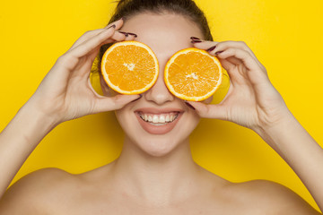 happy young woman posing with slices of oranges on her face on yellow background