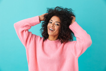 Wall Mural - Colorful portrait of smiling woman in pink shirt posing on camera and touching her dark afro hairdo, isolated over blue background