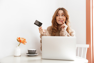 Wall Mural - Photo of adorable brunette woman 20s in sweater thinking and holding credit card, while doing payment transaction in coffee shop