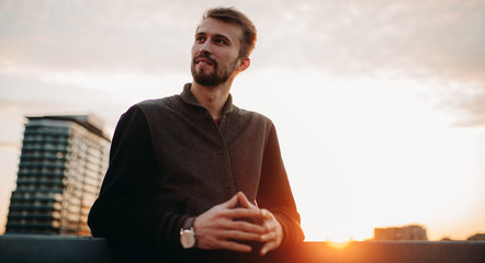 Young man stands and smiles on house roof against background of skyscrapers and sunset.