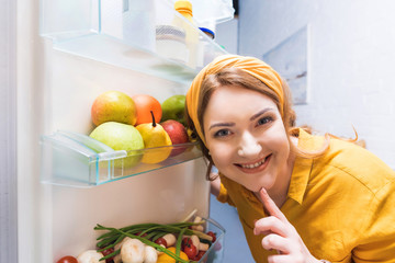 smiling beautiful woman looking at camera near open fridge at kitchen