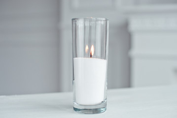 A burning candle in tall glass vase flask standing on the table indoor.