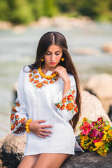 brunette with yellow decorations sits on a stone in a white embroidered tunic