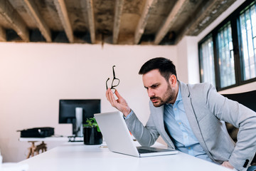 close-up image of a serious business man holding eyeglasess while looking at laptop screen.