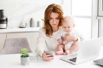 Portrait of young business woman sitting at the table and using her cellphone while holding her cute little baby in hand isolated