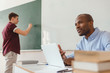 © LIGHTFIELD STUDIOS - High school teenage student writing on chalk board and african american teacher sitting at desk with laptop and explaining