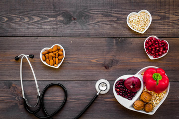 Products good for heart and blood vessels. Vegetables, fruits, nuts in heart shaped bowl near stethoscope on dark wooden background top view copy space