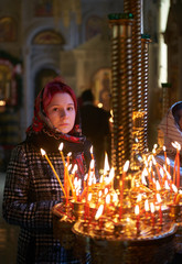 Wall Mural - Praying young woman with candle near pedestal with many other candles in church by her hand to show her faith and esteem to God