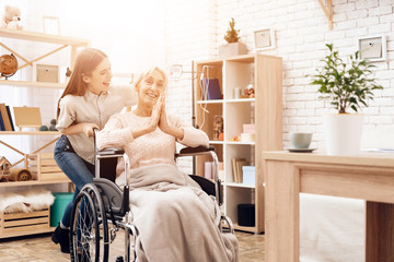 Girl is nursing elderly woman at home. Girl is riding woman in wheelchair. Woman is enjoying herself.