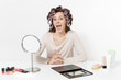 © ViDi Studio - Fun young woman with curlers on hair sitting at table applying makeup with set facial decorative cosmetics isolated on white background. Beauty female fashion lifestyle concept. Area with copy space.