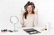 © ViDi Studio - Fun woman with curlers horns gesture depicting heavy metal rock sign at table applying makeup with set facial decorative cosmetics isolated on white background. Beauty female fashion lifestyle concept