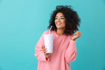 Wall Mural - Portrait of adorable woman 20s with afro hairdo looking aside while holding soda beverage in paper cup, isolated over blue background