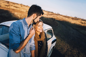 Wall Mural - Young beautiful couple hugging each other, sitting on a small white car in beautiful evening light. Stylish guy with a beard and blond girl laughing