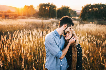 handsome guy with a beard in a blue denim shirt gentle hugs, hand holding and kissing a girl with blond hair in a blue dress and yellow scarf in a field at sunset. stylish couple