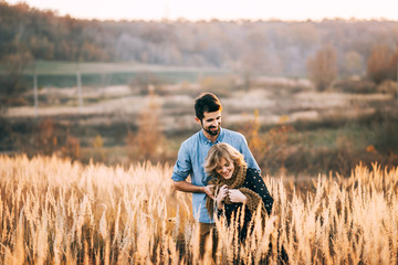 Wall Mural - handsome guy with a beard in a blue denim shirt gentle hugs, hand holding and kissing a girl with blond hair in a blue dress and yellow scarf in a field at sunset. stylish couple