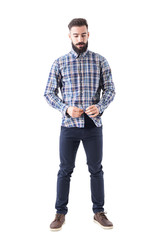 Wall Mural - Relaxed cool bearded hipster buttoning plaid checkered shirt looking down. Full body isolated on white background. 