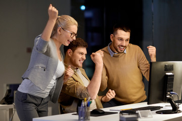 business, success and teamwork concept - happy coworkers with computer celebrating victory at night 