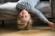 Cute funny boy lying upside down on sofa looking at camera, smiling playful preschool child having fun at home on couch, head shot portrait of happy blue-eyed kid with light hair playing at home