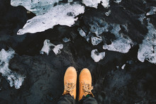 Traveler In Hiking Boots Stand On Black Sand. Iceland. Top View