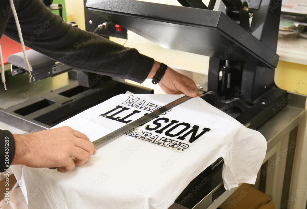 Young man printing on t-shirt at workshop - obrazy, fototapety, plakaty 