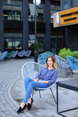 Wall Mural - Cute woman sitting in chair with building background and wearing jeans. concept of fashion look and urban life.