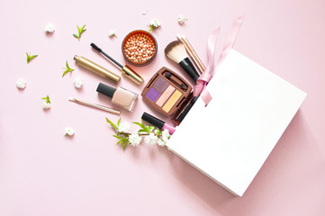 Various cosmetic products eye shadow powder lip gloss blush mascara brush for makeup and white spring flowers in a white gift bag on a pink pastel background top view with copy space