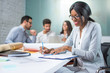 © Bojan - Smiling young business woman taking notes to notebook while sitting in office with colleagues in the background.