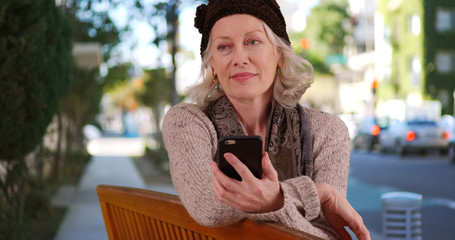 Wall Mural - Elderly white woman sitting outside on public bench with smartphone in hand