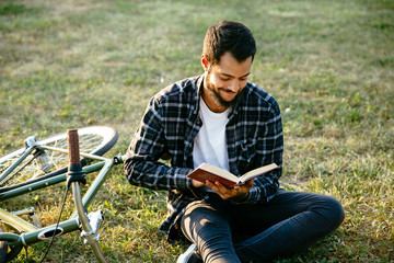 Attractive smiling bearded man reading interesting book, spending time with pleasure while sitting on the grass near the bike. Leisure concept.