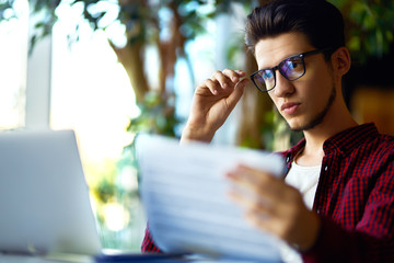 Smiling young hipster man in glasses with laptop on the table. Programmer, web developer, designer working at office comparing mobile and desktop website versions.
