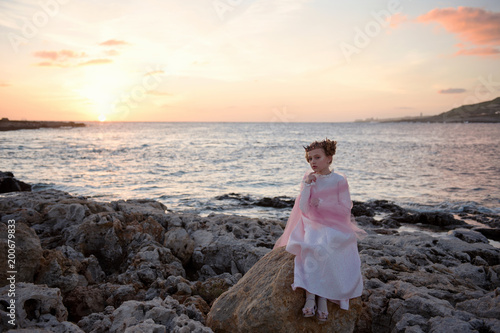 Fotografia, Obraz pensive sad princess girl in a pink dress and a diadem is sitting on a rock on t