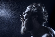 A Bearded Man Angrily Screams Into A Spray Of Water Against A Black Background. Toned Image.
