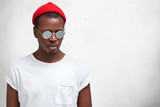 Fototapeta  - Isolated shot of handsome stylish black man in fashionable shades and hat, looks thoughtfully down, poses against white concrete background with copy space for your advertisment or promotion