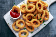 © grinchh - Fried Onion Rings with Ketchup on white cutting board.