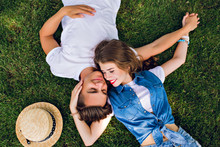 Romantic Couple Of Young People Lying On Grass In Park. They Lay On The Shoulders Of Each Other And Hold Hands Together. They Look Happy. View From Above.