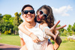 © Look! - Portrait of cute couple having fun in park. Pretty girl with long curly hair is riding on back of handsome guy. They wear sunglasses and smile to camera.