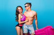 © Look! - Young couple of hot guy with a bare torso hugging pretty girl with long curly hair in swimsuit on blue  background in studio. They wear sunglasses and he holds pink mattress.