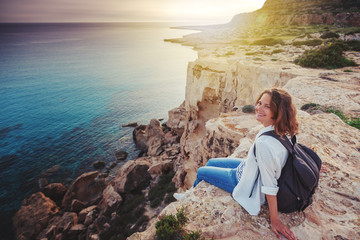 Wall Mural - A stylish young woman traveler watches a beautiful sunset on the rocks on the beach, Cyprus, Cape Greco, a popular destination for summer travel in Europe