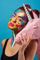 Pop art portrait of model wearing black opened top. Girl has saturated make up with bright geometrical figures and fancy hairdress. Posing on blue background with pink paper unicorn's head.